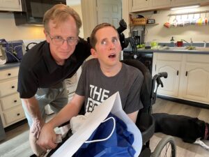 Dad Paul bends down next to son, Sam in his wheelchair. Sam is a TBI Survivor and is holding his UI art package that just came in the mail.